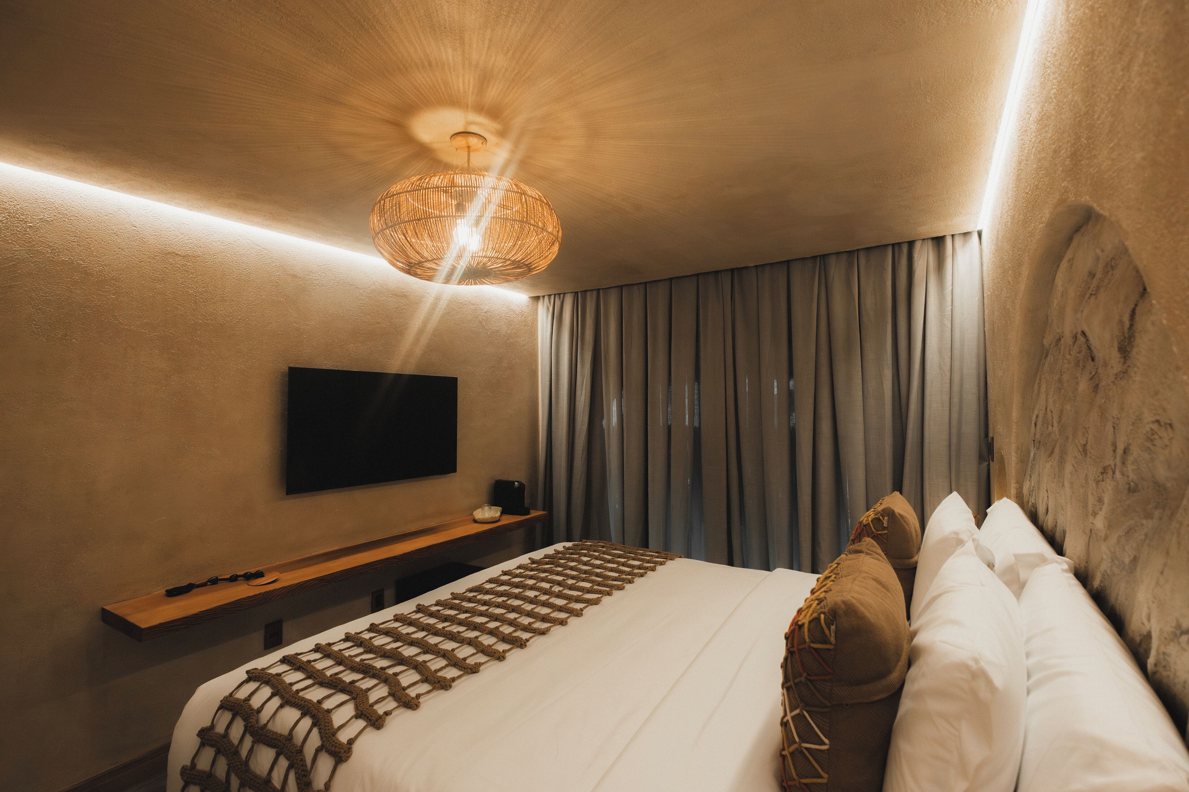 Quarto de hotel com cama grande, televisão de tela plana na parede, mesa de cabeceira de madeira e luminária de tecido.