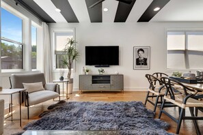 A modern living room with a large flat-screen TV, grey sofa, dining table with chairs, and a blue shag rug.