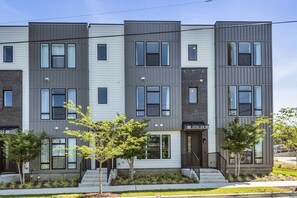 A multi-story residential building with a modern architectural design, featuring a combination of grey and white exterior panels, multiple windows per floor, and a small front garden with young trees.