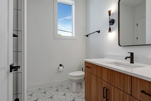 Un baño moderno con inodoro blanco, tocador de madera, un espejo grande y suelo con estampado geométrico.