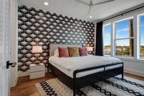 Un dormitorio con papel tapiz estampado, una cama con numerosas almohadas, lámparas de noche y vistas amplias a través de grandes ventanales.