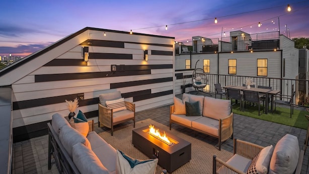 A rooftop patio with outdoor furniture, a fire pit, and string lights against a twilight sky.