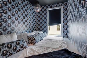 Une chambre avec un papier peint à motifs, un lit avec une literie blanche et une fenêtre avec un store foncé.