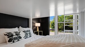 A bedroom with a large bed, a nightstand, a lamp, and a view of greenery through a window.