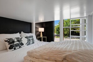 Une chambre à coucher avec un grand lit, une table de chevet, une lampe et une vue sur la verdure par la fenêtre.