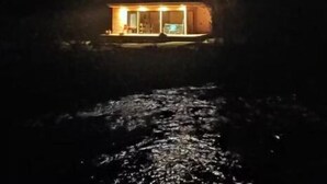 A cabin with a glass door entrance, a visible interior with a seating area, and a river in the foreground. - Untamed Refuge in Rio Puelo (Arrayan) (Los Lagos)