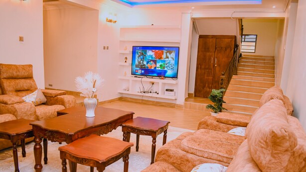 A living room with a flat-screen TV, wooden furniture, and a staircase leading to another level. - Lux Suites Lions Den Holiday Homes (Nakuru)