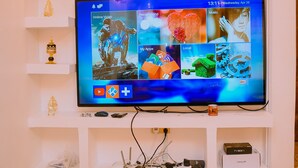 A flat-screen TV mounted on a white wall with a shelf below displaying decorative items and a TV box. - Lux Suites Lions Den Holiday Homes (Nakuru)
