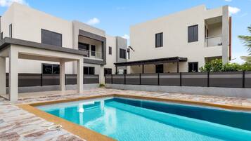 A modern pool area with a rectangular pool, a covered seating area, and a multi-story building with balconies.