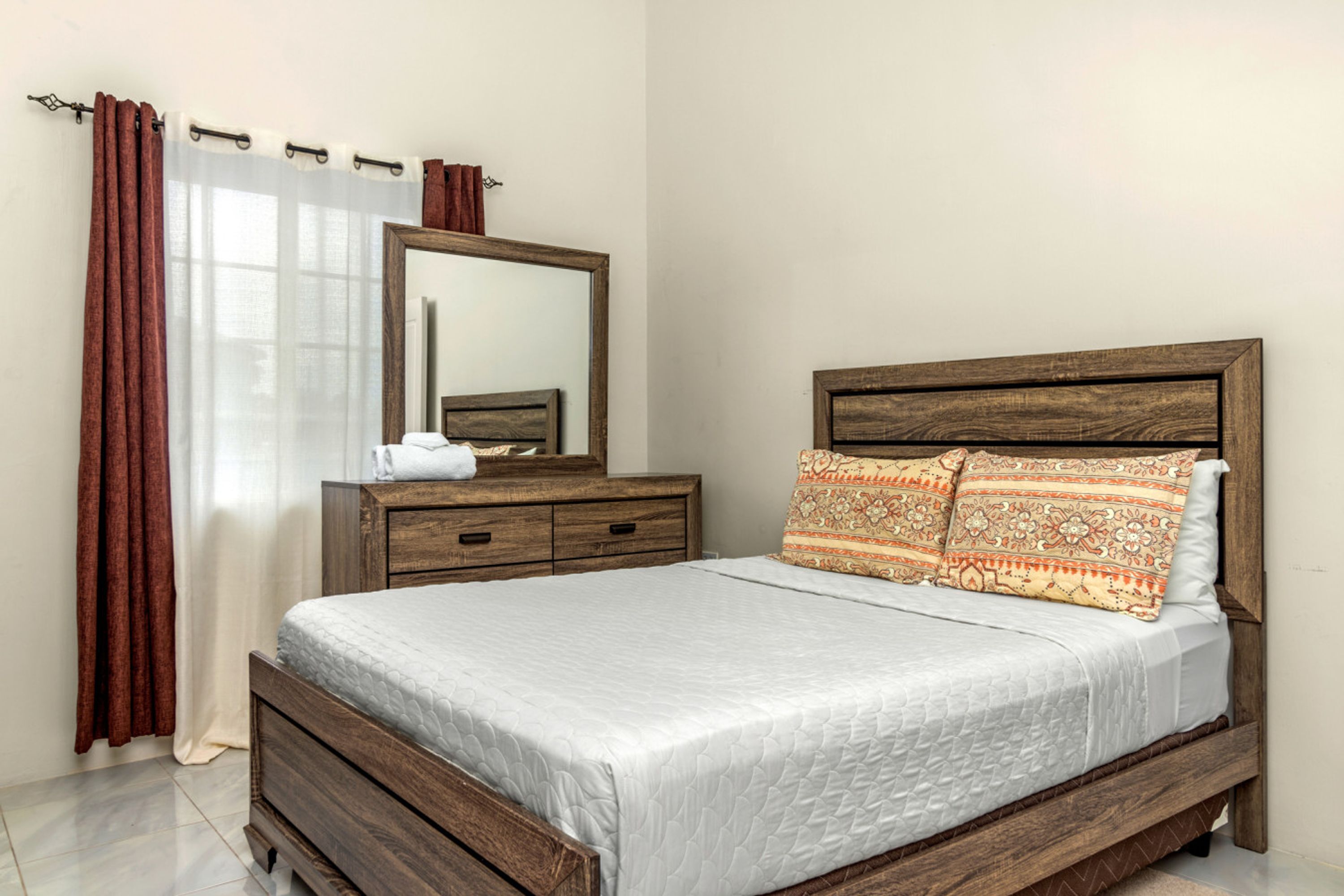 A bedroom with a wooden bed frame, a nightstand, a mirror, and a window with curtains.