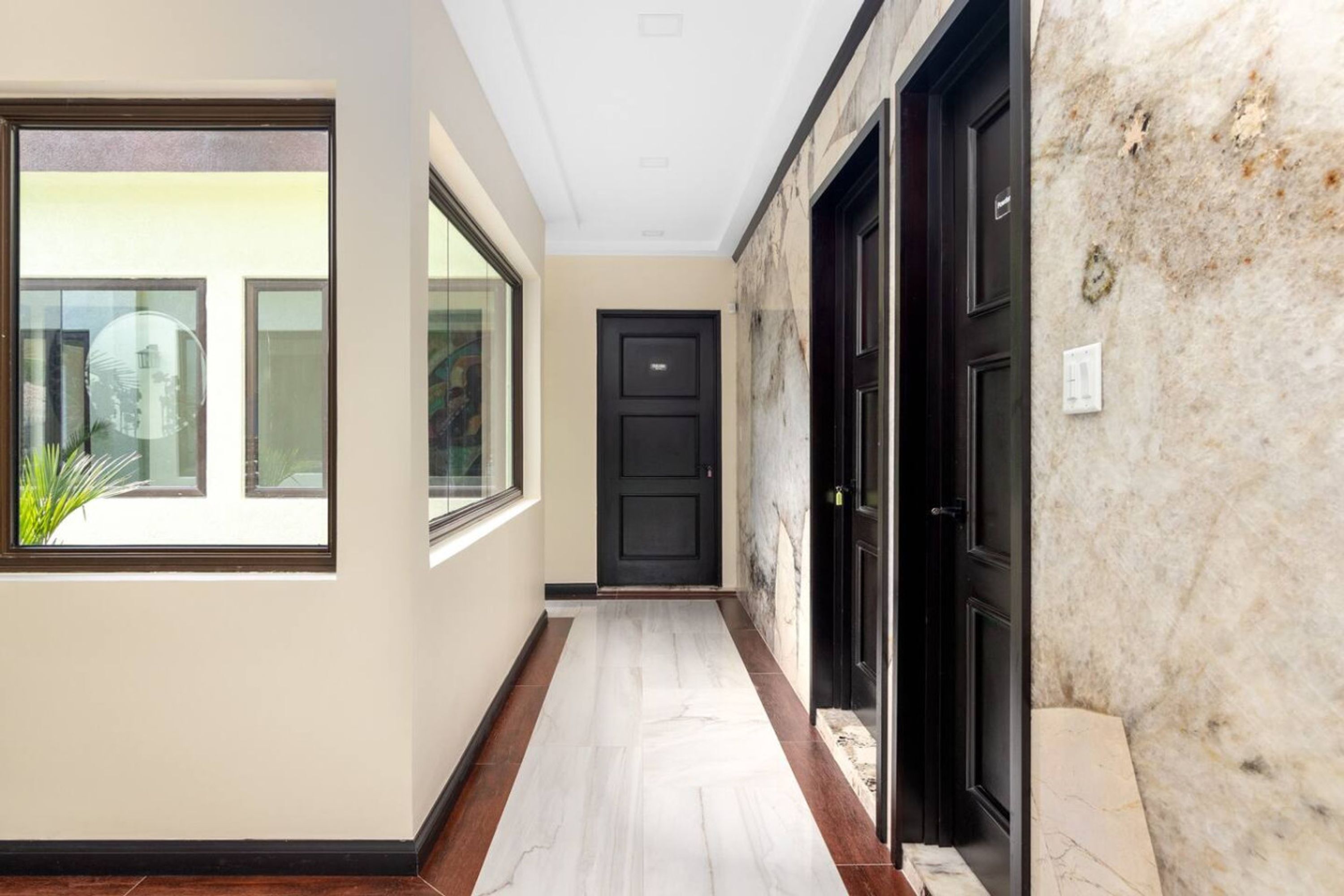 A hallway with a marble wall, a black door, and a window with a view of greenery.