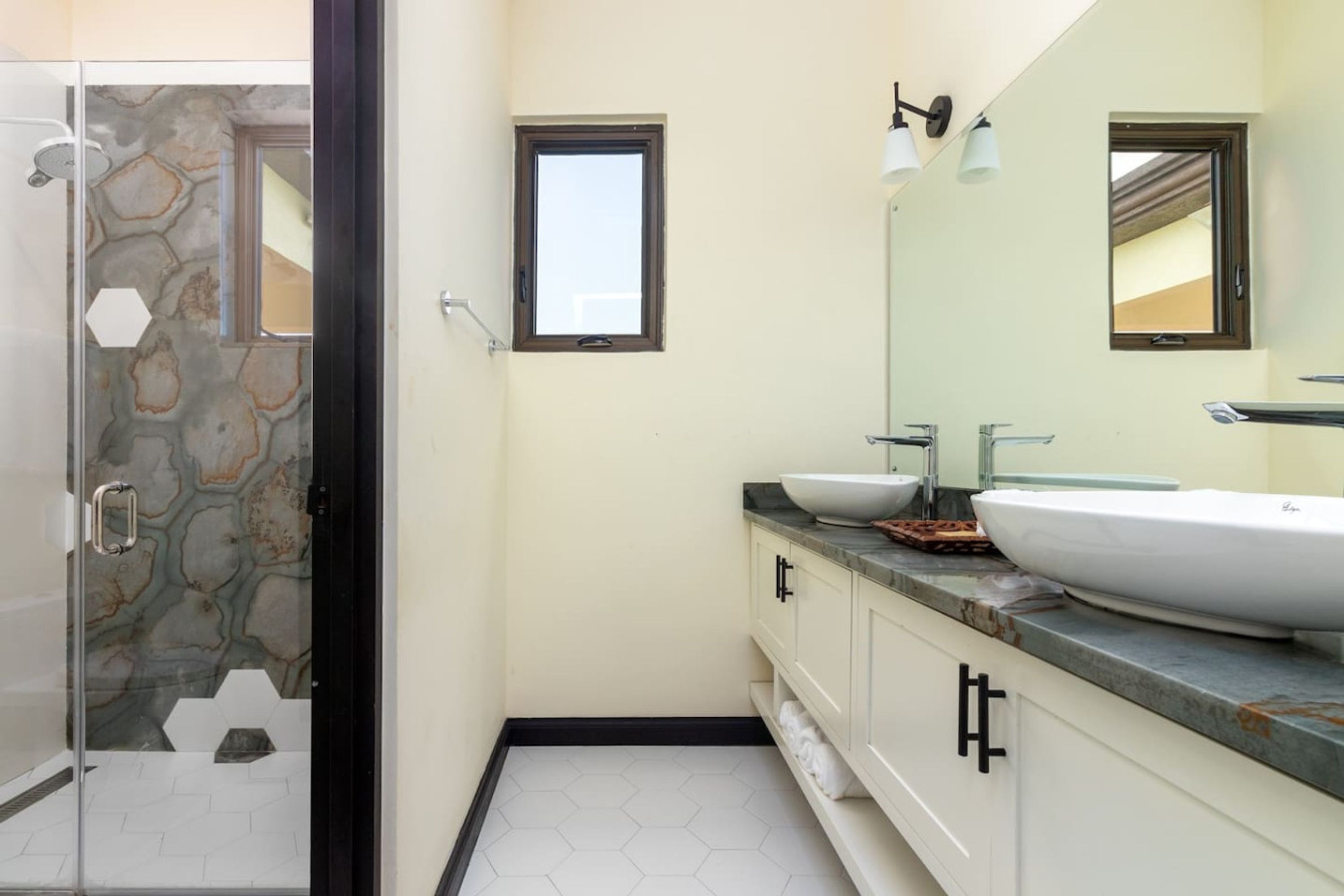 Un baño con cabina de ducha de vidrio, dos lavamanos con encimeras de mármol y un espejo grande sobre el mueble.