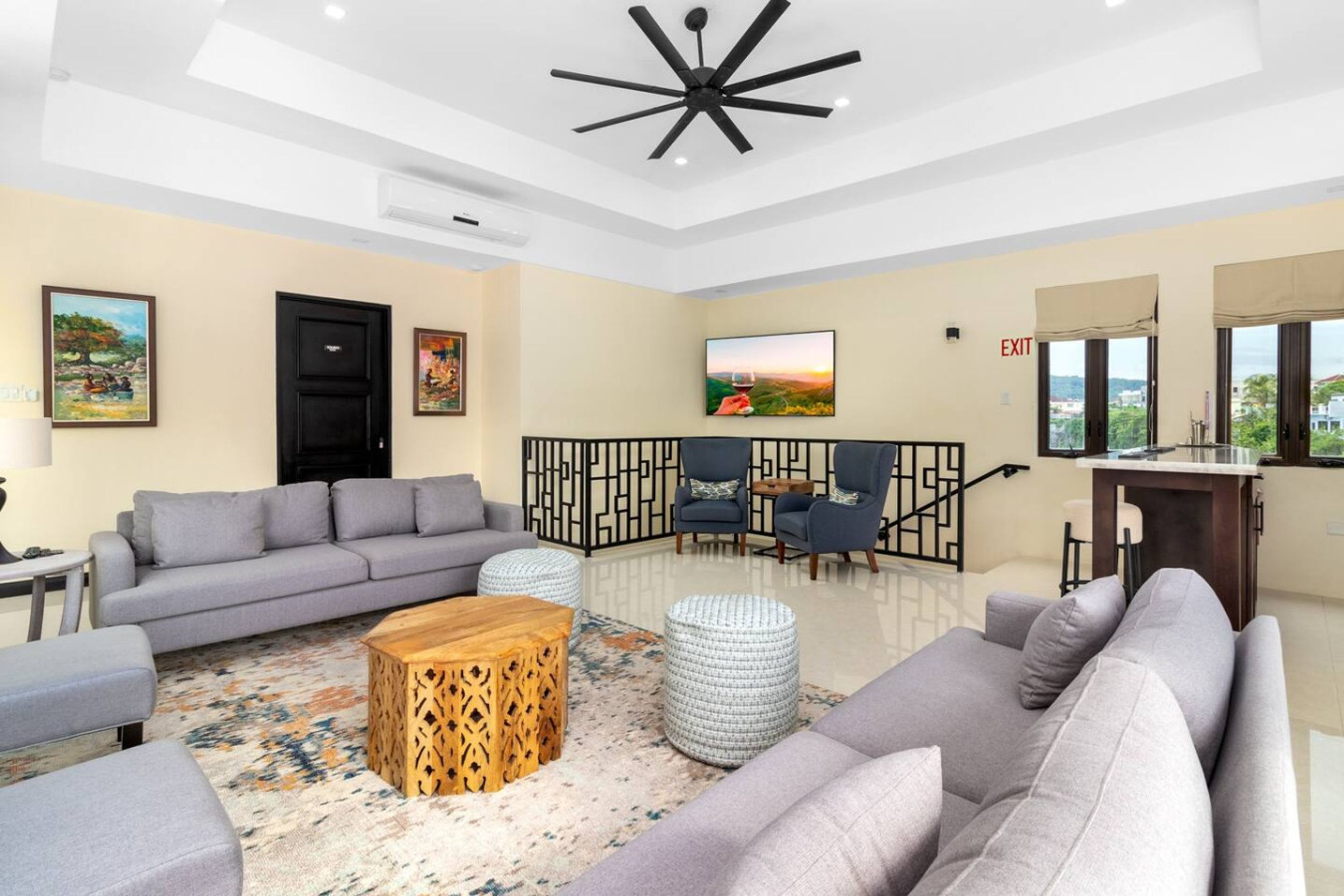 A modern living room with a grey sofa set, a wooden coffee table, and a patterned rug. There is a black door, a TV mounted on the wall, and a view of the outside through large windows.