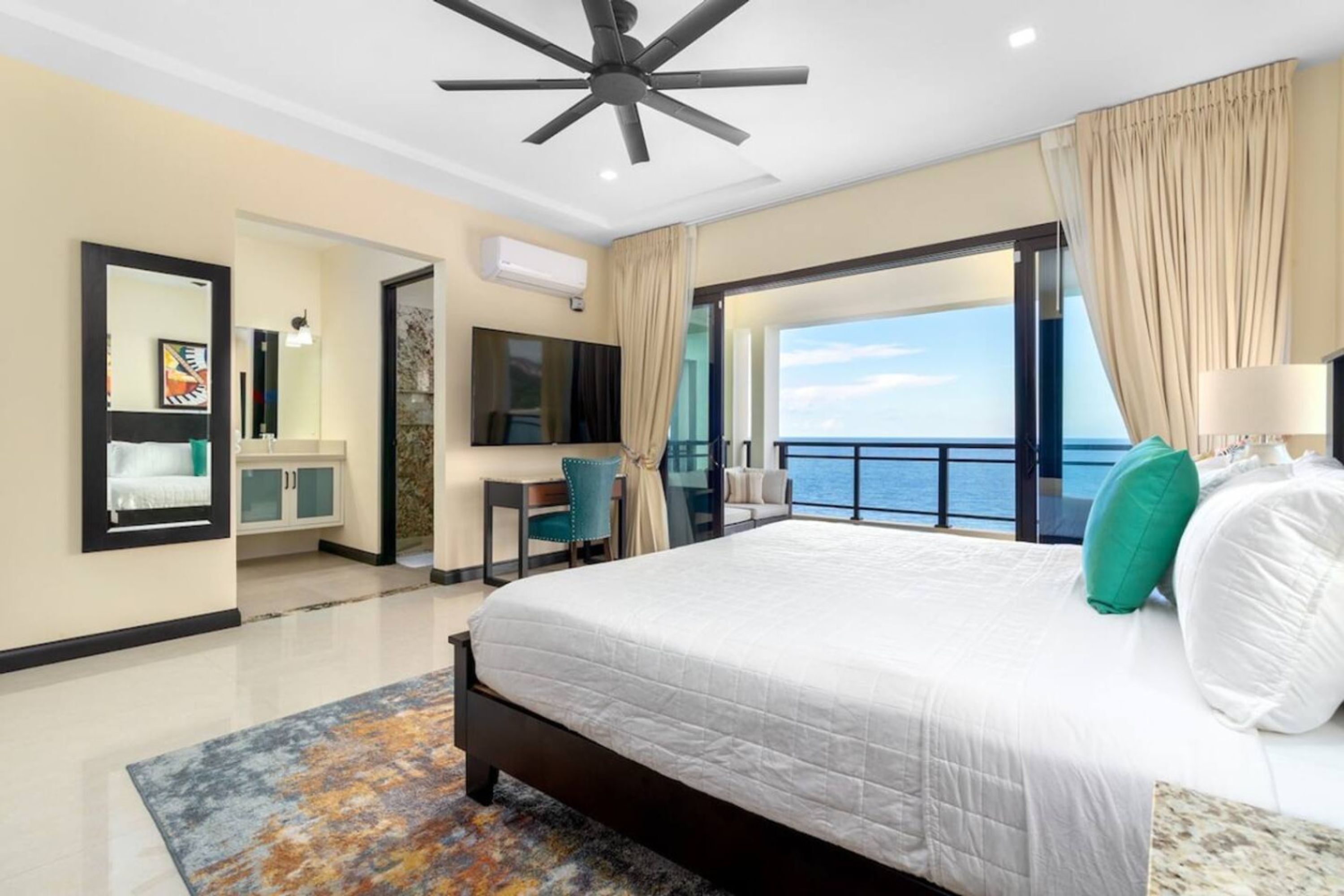 A modern hotel room with a large bed, a desk, a TV, and a balcony with a view of the sea.