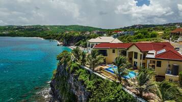 A coastal property with a swimming pool, a red-roofed building, and a view of the sea and hills.