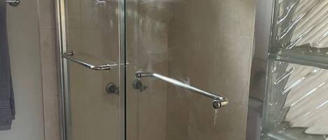 A glass-enclosed shower with a metal handrail, a window with a geometric pattern, and a tiled floor.
