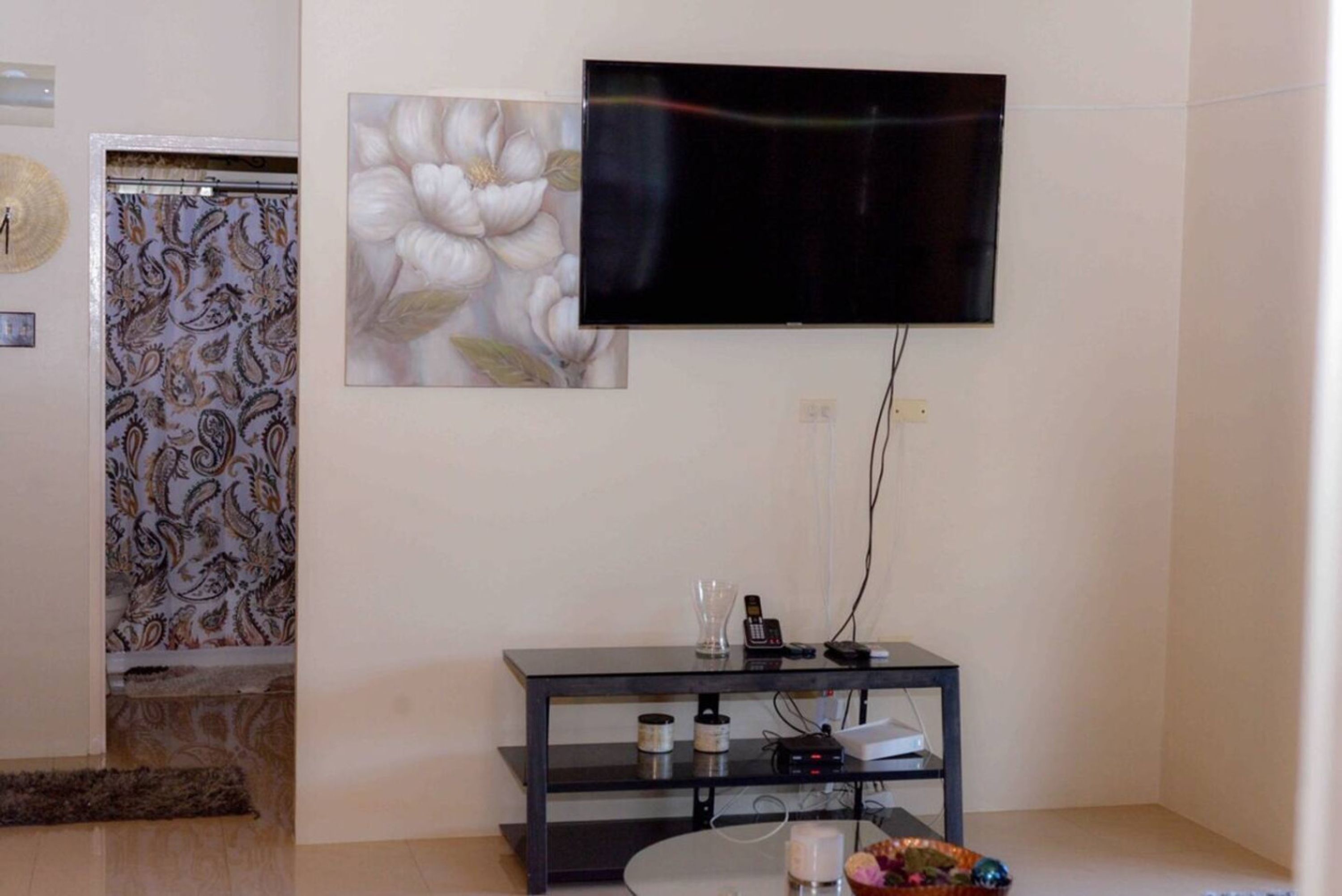 A living room with a flat-screen TV mounted on the wall, a small black TV stand with electronic devices, and a decorative flower painting.