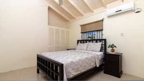 Ein Schlafzimmer mit Bett, Nachttisch, Topfpflanze, einem Fenster mit Jalousien und einer Klimaanlage.
