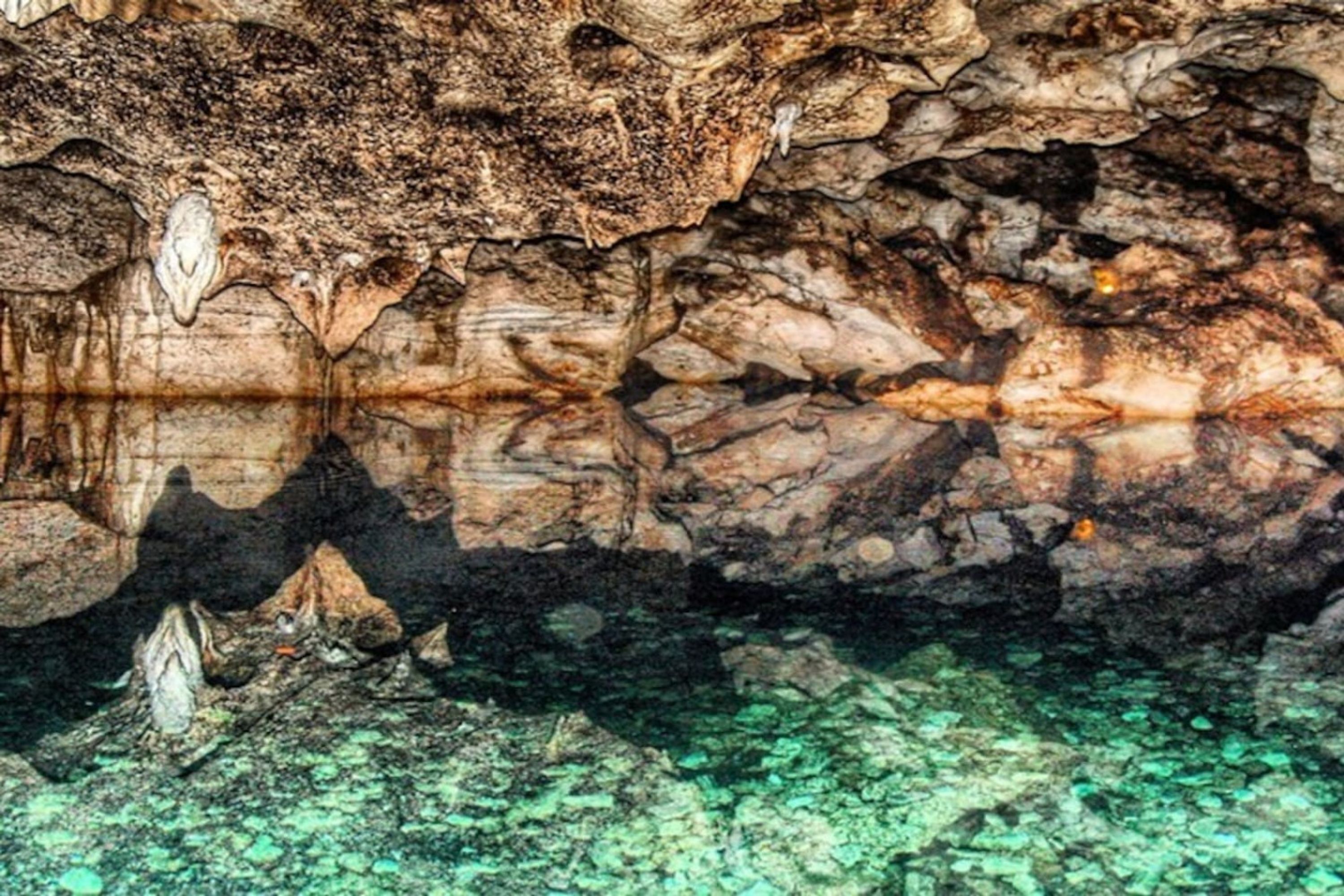 Interior de cueva con estalactitas y estalagmitas, agua cristalina y suelo rocoso.