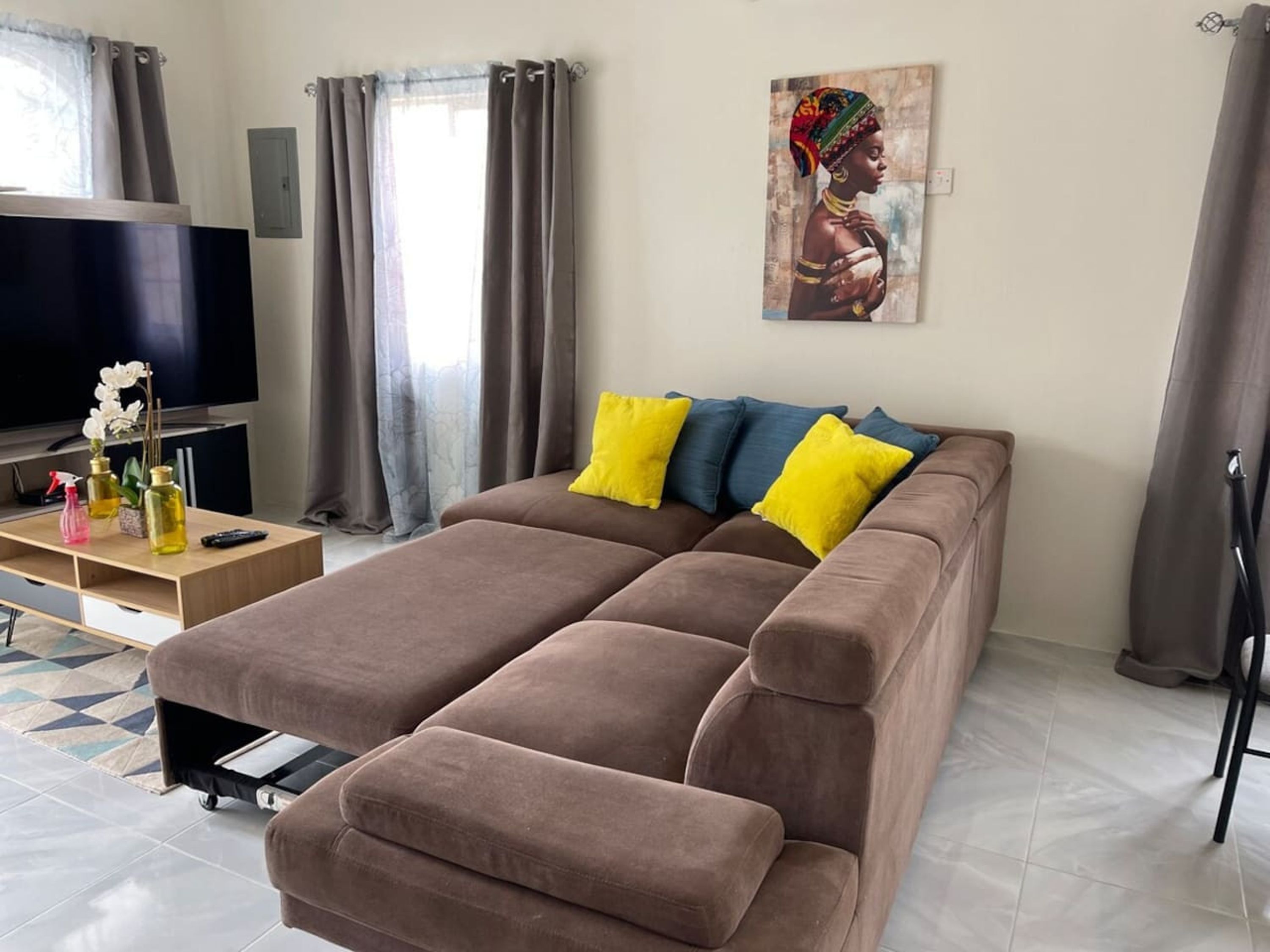 A modern living room with a brown sofa, two yellow and one blue pillow, a flat-screen TV, a wooden coffee table, and a painting on the wall.