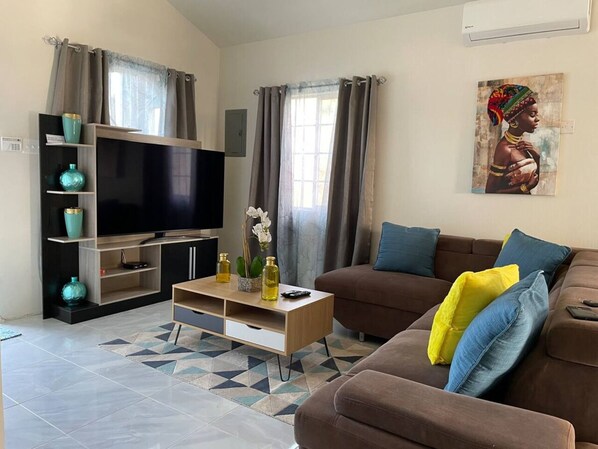 Una sala de estar con un televisor de pantalla plana, un sofá marrón con cojines coloridos, una mesa de centro de madera y un tapiz estampado.