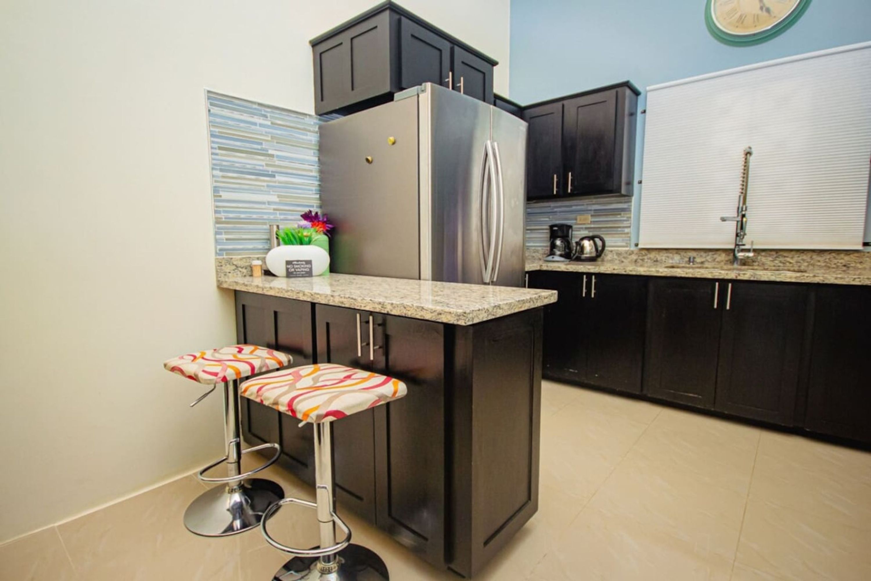 Une cuisine avec des meubles de couleur sombre, un réfrigérateur en acier inoxydable, un tabouret de bar et une horloge au mur.