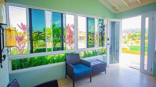 A room with large windows, a wicker sofa, and a view of a green landscape. - Adorable Beach House in Drax Hall Gated Pool Gym (St. Ann's Bay)