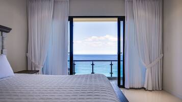 A bedroom with a large bed, a view of the ocean through an open door, and a balcony with a railing.