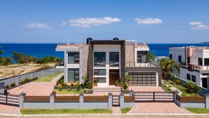 A modern two-story house with a large garage, a well-maintained garden, and a view of the ocean. - Luxury Oceanfront Villa Fully Staffed Pool Jacuzzi Movie Theatre (Tower Isle)