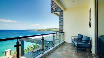 A balcony with a view of the ocean and a stone wall. There are two chairs and a table on the balcony.
