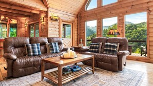 A cozy cabin interior with two leather sofas, a wooden coffee table, and large windows offering a view of the forest.