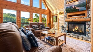 A log cabin interior with a stone fireplace, wooden furniture, and large windows offering a view of the forest.