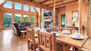 A wooden cabin with a dining area, fireplace, and large windows overlooking a forested landscape.