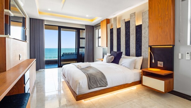 A modern bedroom with a large bed, a wooden bedside table, a built-in shelf, and a view of the ocean.