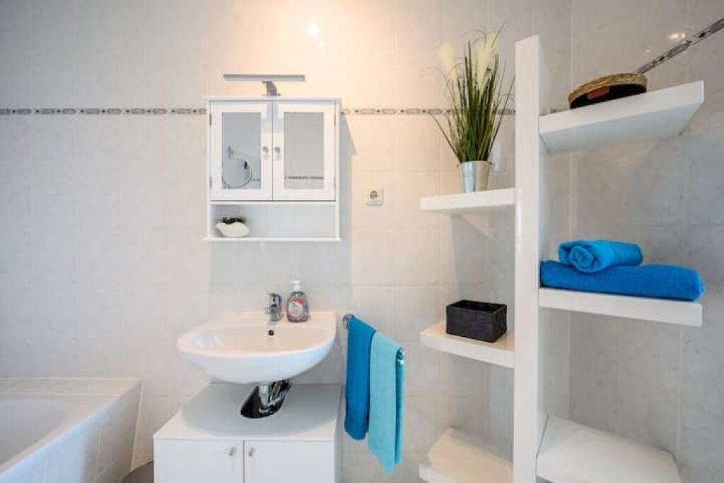 Een badkamer met een witte wastafel, een spiegelkast, een handdoekrek en een plank met opgevouwen handdoeken.