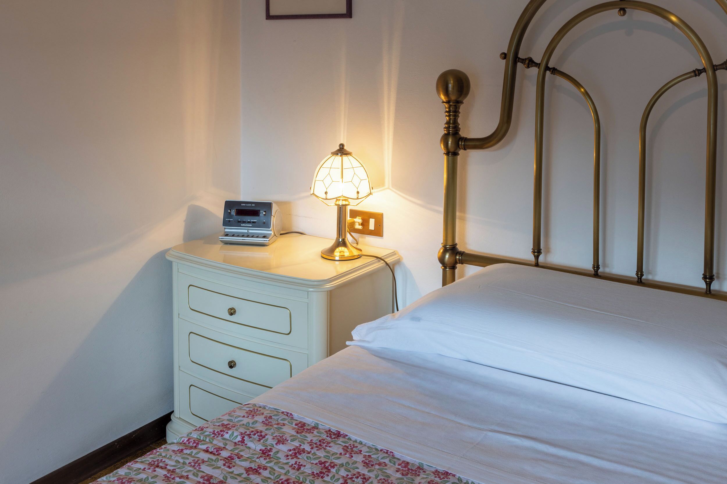 A neatly made bed with a floral-patterned bedspread, a bedside table with a lamp, and a wall-mounted radio.
