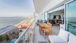 A balcony with a view of the beach and ocean, a dining area with wooden furniture, and a modern interior with a large window. - Beachfront Peninsula PV 8th Floor (Puerto Vallarta)