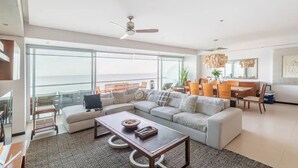 A modern living room with a large sofa, a coffee table, and a dining area with wooden chairs and a table. There is a balcony with a view of the ocean. - Beachfront Peninsula PV 8th Floor (Puerto Vallarta)