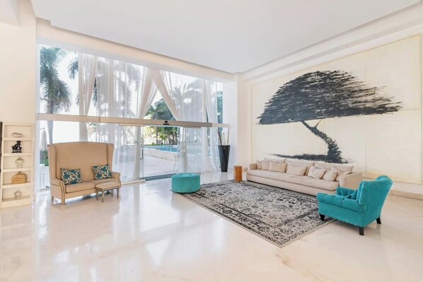 A spacious living room with a large window, a beige sofa, a teal armchair, and a black and white patterned rug.