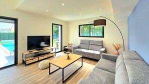 Una sala de estar moderna con un televisor de pantalla plana, sofás grises, una mesa de centro de madera y un ventanal que ofrece vistas al exterior.