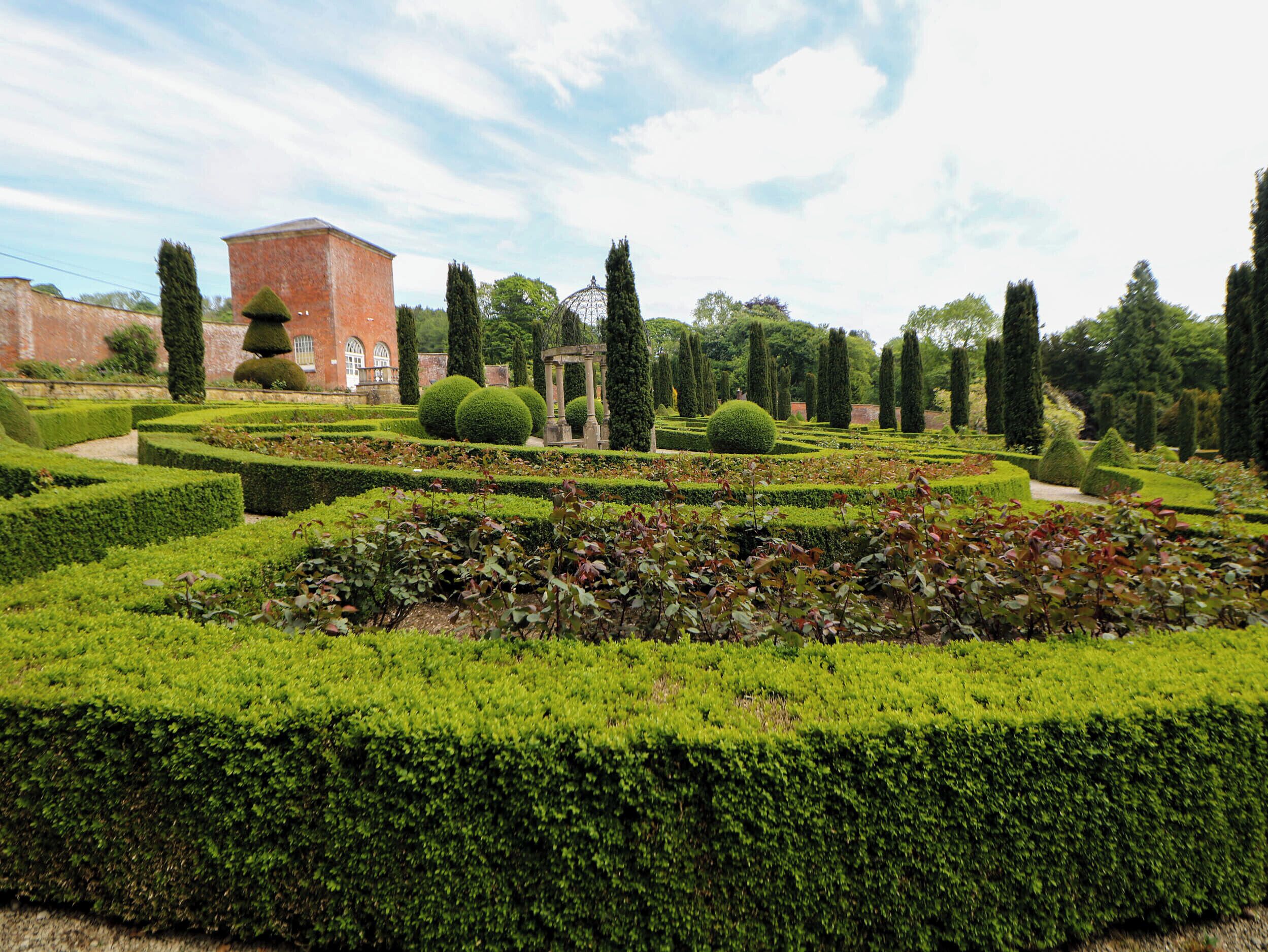 Un jardin à la française, avec des haies aux formes géométriques, une fontaine centrale et un bâtiment de style classique en arrière-plan.