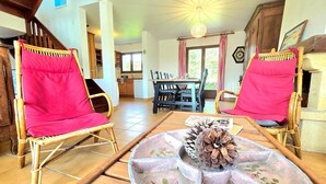Ein gemütliches Wohnzimmer mit zwei Korbstühlen, einem Holztisch mit einer Kiefernzapfen-Dekoration und einem Essbereich im Hintergrund.