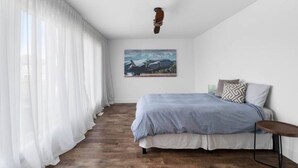 Un dormitorio con una cama, un cuadro en la pared y una ventana con cortinas ligeras.