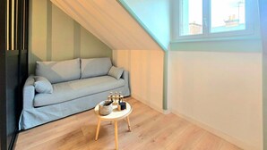 A small, cozy room with a sloped ceiling, a light grey sofa, a small wooden table with a basket and a candle holder, and a window with a view of buildings. - A pas de tout, historic center, train, parking (Vannes)