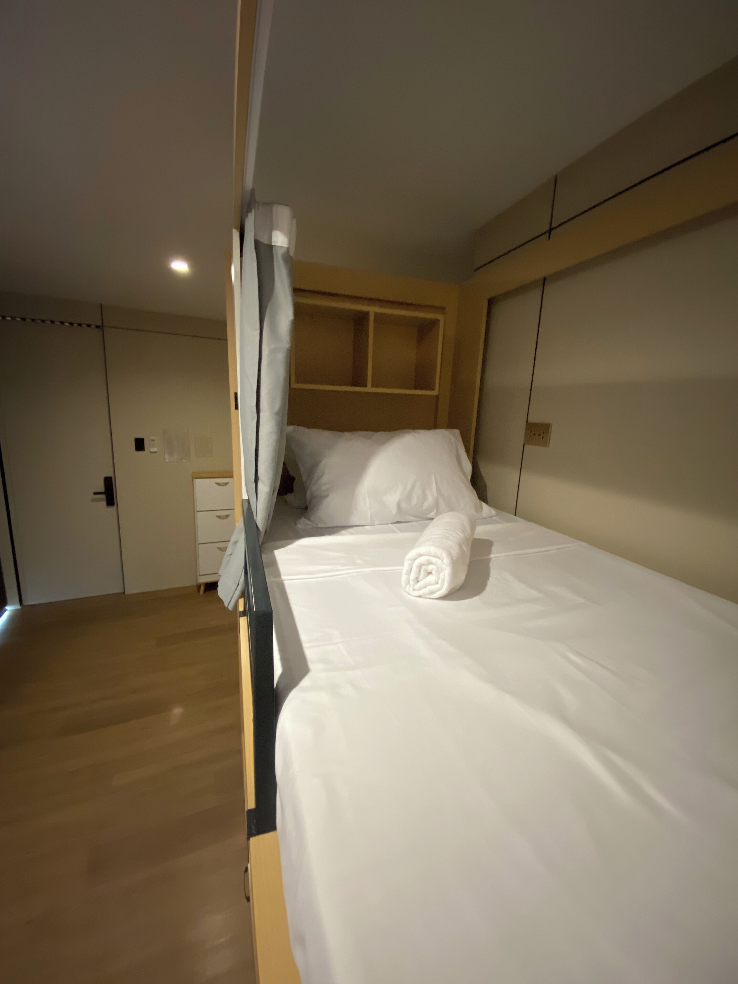 A single bed with white linens and a rolled towel, a wooden headboard, and a white curtain.