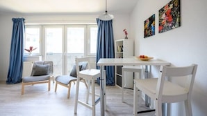 Une pièce lumineuse avec une table blanche, deux chaises blanches, un rideau bleu et un tableau accroché au mur.