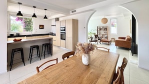 Une cuisine moderne dotée d’un îlot central, de luminaires suspendus noirs et d’un coin repas avec une table et des chaises en bois.