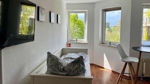 A small room with a bed, a TV mounted on the wall, and two windows providing views of greenery.