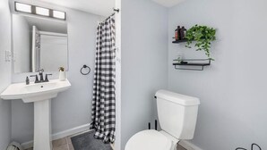 A bathroom with a white sink, a toilet, a checkered shower curtain, a mirror with lights, and a shelf with plants.