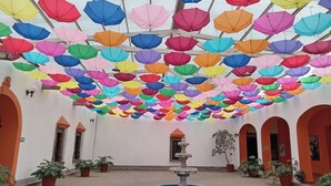 A courtyard with a fountain, surrounded by potted plants, and colorful umbrellas hanging from the ceiling. - Hotel Hacienda La Muralla (Amealco)